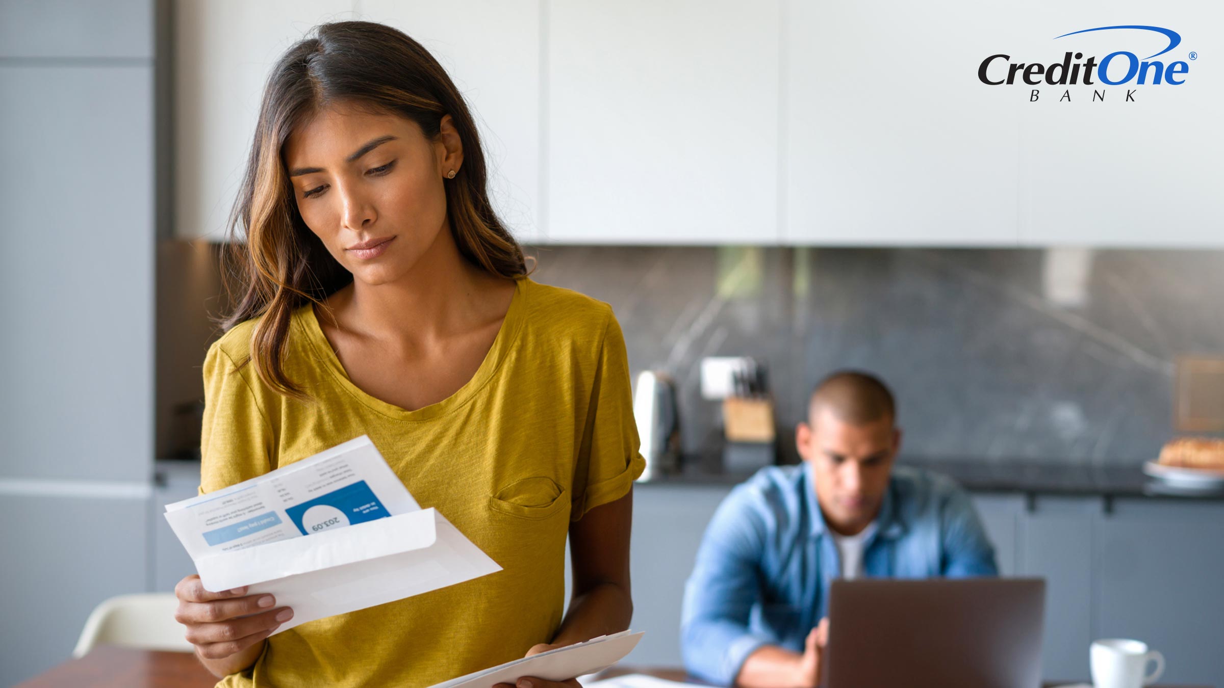 A man works on his laptop in the kitchen as his wife looks at a collections letter with a concerned expression. She may be worried about how to remove it from her credit report.